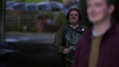 Lorcan wears a forest green hooded jumper outside and smiles. A blurred figure in the foreground s=covers the image of Lorcan