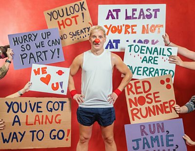 Jamie Laing stood in running shorts, a vest and a red nose with his hands on his hips.  Behind him are arms holding various homemade cardboard signs of support 