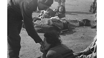 Black and white photograph of man dressed in army uniform reaching down to a woman. The woman clasps his hand to her cheek. She is wrapped in a blanket and large jacket