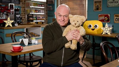 Ross Kemp hugs a fluffy teddy bear to camera. He is sitting in a cafe set with a teacup and saucer on the table beside him. A yellow CBeebies 'bug' with a smiley face sits on the table behind him