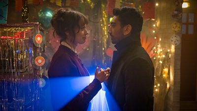 Aimee Lou Wood and Nabhaan Rizwan stand opposite one another in the light from a projector. A darkened room around them is illuminated by the projector and fairy lights, and is decorated with homemade crafts. 