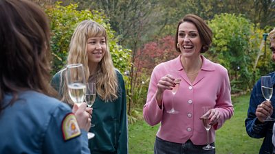 Suranne Jones stands beside other characters in a garden. They all hold a champagne glass as they chat. 