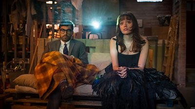 Nabhaan Rizwan and Aimee Lou Wood sit beside each other on a make-shift sofa made of blankets and wooden pallets. A film projector shines a light between them as they watch the projection. 