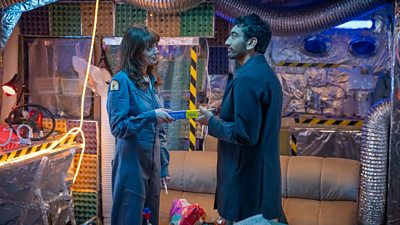 Aimee Lou Wood and Nabhaan Rizwan stand opposite one another. They each hold onto the same tube of kitchen foil with an amused expression. Furniture in the background is wrapped in foil and illuminated by golden, glowing lights. Popcorn and snacks are visible on the table beside them. 