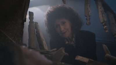 A still of Anita Dobson in her role as Angie Watts on the set of EastEnders. She sits on the staircase of a house and looks down through a broken bannister. 