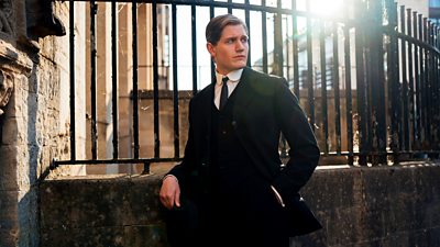 Adam Hugill as Mac in Agatha Christie’s Towards Zero. Wearing a black suit that is tailored in a style linked to the early 1900s. He leans against a metal railing outdoors. The sun shines through the railings behind him, creating a lens flare effect on the image. 