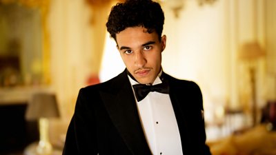 Khalil Gharbia as Louis Morel in Agatha Christie’s Towards Zero. Wearing a tuxedo, he stands in a grand room, decorated with warm colours, lamps and a mirror with gold detailing. 