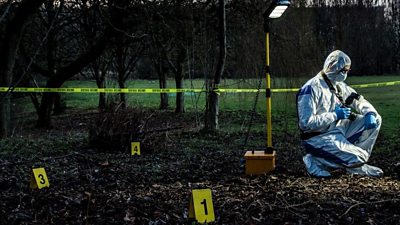 Figure in a hazmat suit crouches in grass surrounded by plastic markers and flood light.