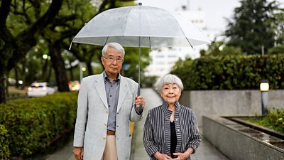 Hiroshi Shimizu and Keiko Shimizu stand under an umbrella smiling