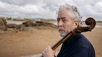 Neil Martin stood on a sandy beach holding a musical instrument of the string kind, only the handle of which, is in shot