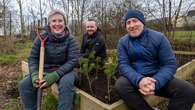 A female and two males sat on a raised flower bed holding gardening tools and smiling. They are dressed warmly.