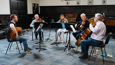 Four male and one female musician sat on chiars in a circle playing music on various string instruments - a guitar, a cello and violins. Behind them in the room is a wooden upright piano and a grand piano