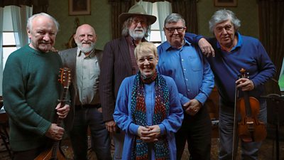 Group shot of six men stood behind a woman. Some of the men are holding instruments including a violin. They are smiling
