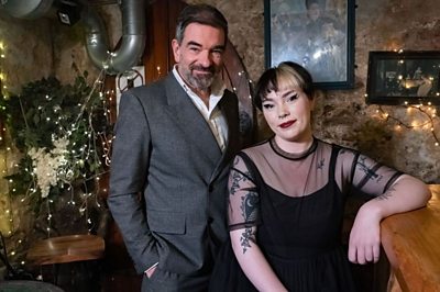 A man in a gret suit stands next to a female in a black lace dress who is sat at what appears to be a bar. There are twinkly fairy lights behind them and some decorative foliage.