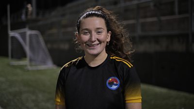 A girl stood on a pitch in front of a goal. She is smiling at the camera. It is night time
