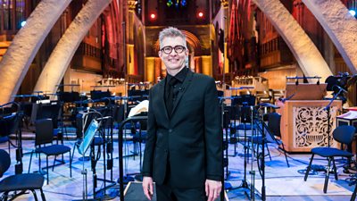 Gareth Malone stood in a beautifully lit Llandaf Cathedral surrounded by an orchestra setup. He wears a black suit