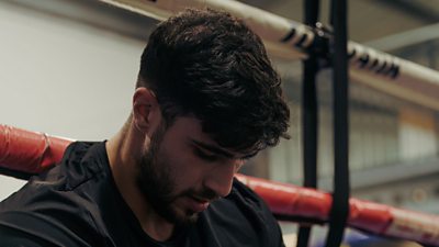 A man in a black t-shirt sits on the edge of boxing ring looking down