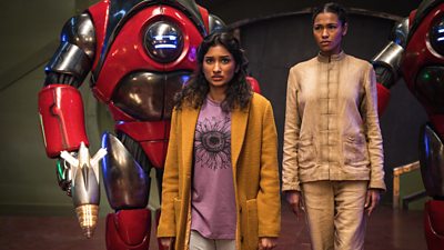 Two woman stand flanked by red robots, who hold futuristic weapons. One woman wears a mustard yellow coat over a pink t-shirt while the other wears a beige top and trousers. They both look concerned.