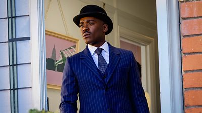 A man in a pinstripe blue suit and a bowler hat looks out the front door of a house with a concerned expression on his face