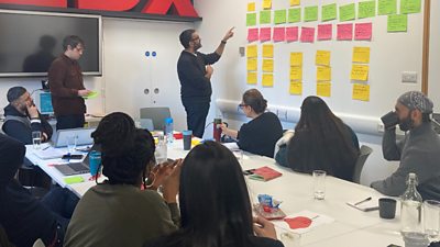 Writers sat around a desk as author AA Dhand (south asian male) stands and points to a wall of post it notes with story beats for Virdee