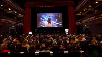 A full audience sitting in stall seating at a theatre with an image of BBC's Virdee on screen over the stage for the premiere screening (Credit: ý / Neil Sherwood)