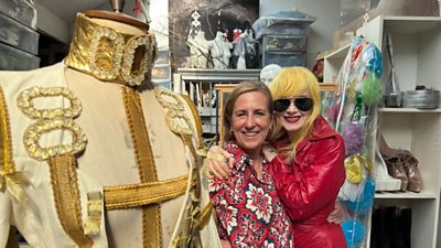 Kirsty Wark et Pam Hogg regardent la caméra s'embrasser devant un grand mannequin vêtu de costumes d'or élaborés. Ils sont à l'intérieur et derrière eux, il y a divers bustes et talons hauts. Pam Hogg porte des lunettes de soleil.