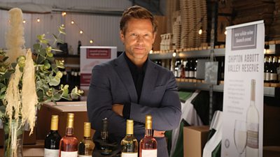 A man in a suit jacket stands smiling with his arms folded behind a counter displaying bottles of wine