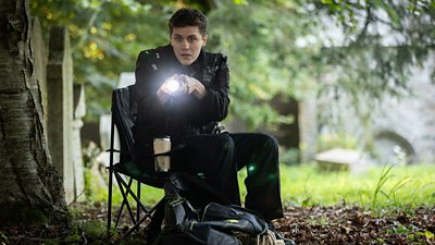 A police officer sitting on a camping chair shines a bright light from a torch at something off screen