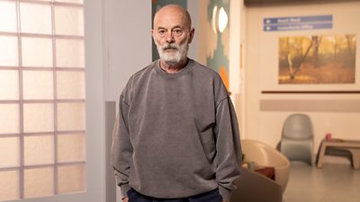 A man in a grey jumper stands in a room, looking straight to camera