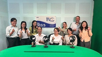 a group of young men and women gather around a green table with microphones - they are smiling - behind then is a sign for a radio station