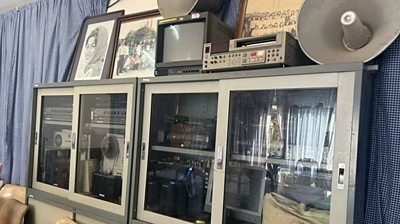 a glass window cabinet is filled with radio equipment and has framed dusty pictures and old equipment on top