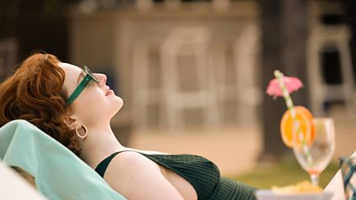 Hannah Onslow in character as Diana Williams. She reclines on a sun lounger outdoors, wearing a green vest top and sunglasses. An empty glass with a straw and garnish is visible on a table in the background. 