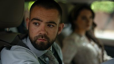 Jamie Phelan in character as Jack McMullen. He’s sat in the driver’s seat of a car, looking at something out the window. The background is blurred, but a woman is visible in the passenger seat beside him. 