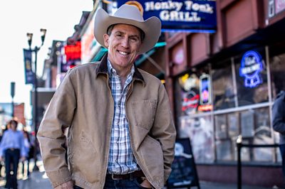 Rob Brydon smiling, wearing a jacket, checked shirt and a stetson stood in front of a sign that reads Honky Tonk Grill