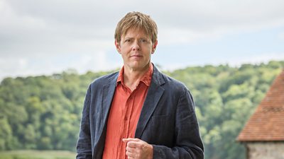 Close-up of Kris Marshall in character as DI Humphrey Goodman. Stood outdoors in the countryside, he looks directly to the camera. Meadows, a forest and a brick building are visible in the background. 