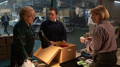 Three women stand around a box on a table in a warehouse discussing something. One wears a police uniform while the others wear civilian clothes