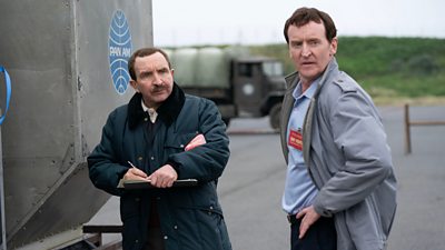 Two men stand outdoors in daylight beside a Pan Am branded silver piece of equipment. A truck is visible in the background. The men wear coats and look concerned by something out of shot. One carries a clipboard and appears to be making notes.