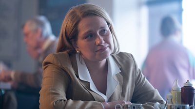A woman in a patterned blouse with a broad white collar sits at a table in a cafe