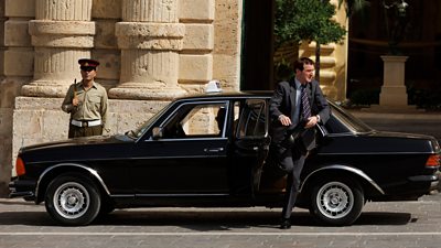 A man gets out of a black car in a sunny place, where a soldier stands guard at a building behind the car