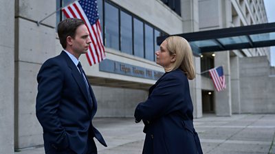 A man a woman stand outside the FBI building, locked in tense conversation. The red and white stripes and blue stars of two United States flags fly from the building.