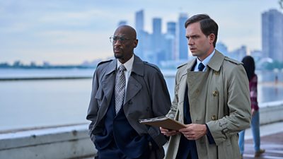 Two men walk along the side of a river with a series of skyscrapers in the distance behind them. One holds a file and both look tense.