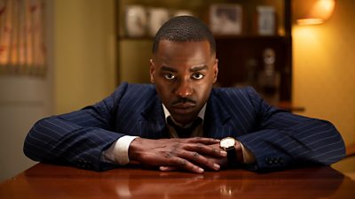A man in a pinstripe suit rests his forearms on a desk with his hands crossed and peers stright down the lens of the camera