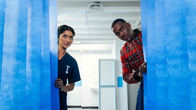 A man and a woman peek from behind blue curtains in a hospital. She wears a nurse's uniform while he wears a tartan style patterned jacket and a kilt