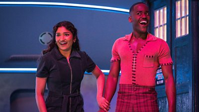A man and a woman stand outside a blue police box on a spaceship, smiling and laughing