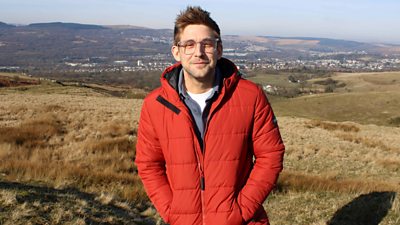 Steffan stood above a hill with the Merthyr Valley behind him. He wears a red puffer coat