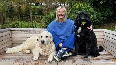 Photo of Jo Whiley sat outdoors on a bench beside two dogs. 
