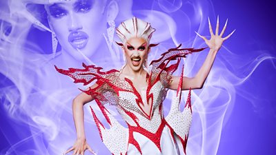 Drag Queen Kyran wearing a flamboyant masterpiece of an outfit resembling flames. His nails are extremely long and he wears dramatic red makeup with a red and white wig