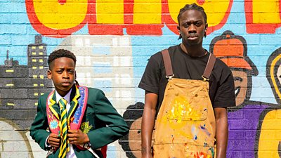 A boy in school uniform and a young man in paint splattered overalls stand in front of a brightly coloured wall