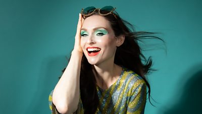 A head and shoulders shot of Sophie Ellis-Bextor, wearing a green and blue glittering dress. She holds one hand to the side of her face, smiling, with a pair of glittering sunglasses on her head.