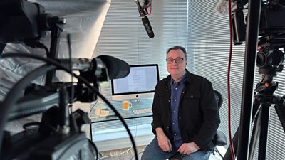 Russell T Davies sits at a glass desk with a computer, cup and telephone. He faces to camera, with a film camera and sound equipment pointed towards him